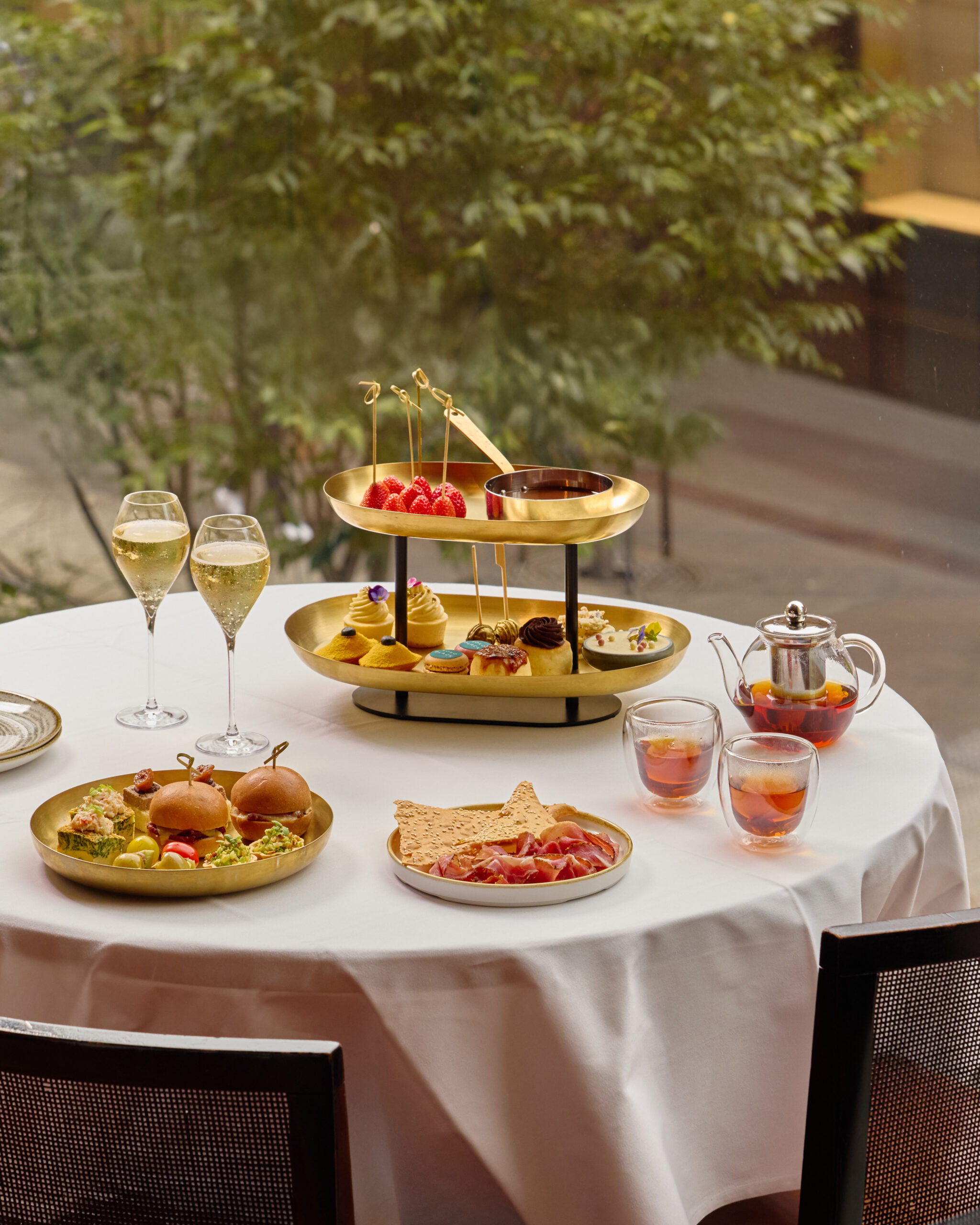 An elegantly set table at Glass Brasserie in Sydney CBD featuring a luxurious high tea with a two-tiered golden stand of handcrafted pastries, delicate desserts, and fresh strawberry skewers dipped in rich chocolate sauce. The spread includes gourmet savoury bites like mini sliders, smoked salmon canapés, and cured meats with crispbread, paired with premium tea served in glass teapots and cups.